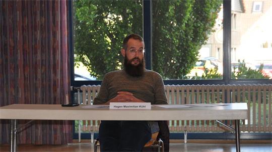 Mann mit Bart und Brille sitzt an einem Tisch mit Namensschild vor großen Fenstern und grünen Bäumen im Hintergrund