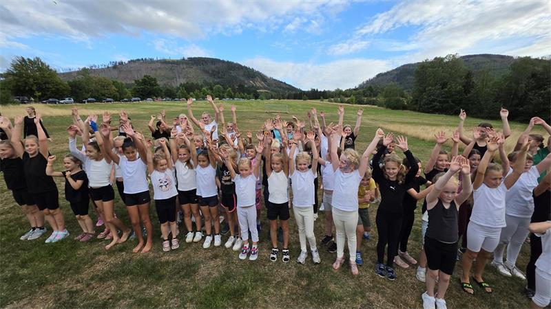 Große Gruppe von Kindern und Jugendlichen steht auf einer Wiese vor bewaldeten Hügeln und hebt die Arme in die Luft.