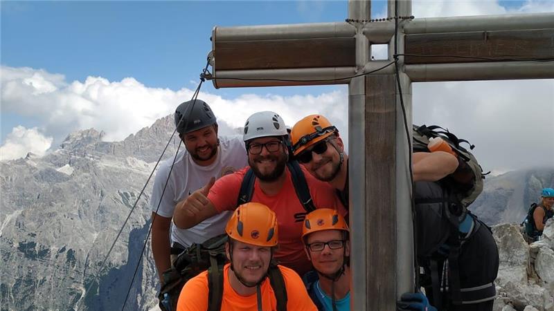 Gruppe von fünf Bergsteigern mit Helmen und Rucksäcken steht an einem großen Holzkreuz auf einem Berggipfel mit Bergpanorama im Hintergrund