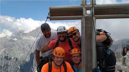Gruppe von fünf Bergsteigern mit Helmen und Rucksäcken steht an einem großen Holzkreuz auf einem Berggipfel mit Bergpanorama im Hintergrund