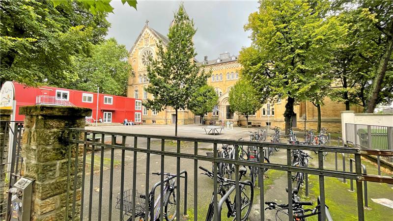 Blick durch ein schwarzes Metallgitter auf einen Innenhof mit mehreren Fahrrädern, Bäumen und einem historischen Backsteingebäude im Hintergrund
