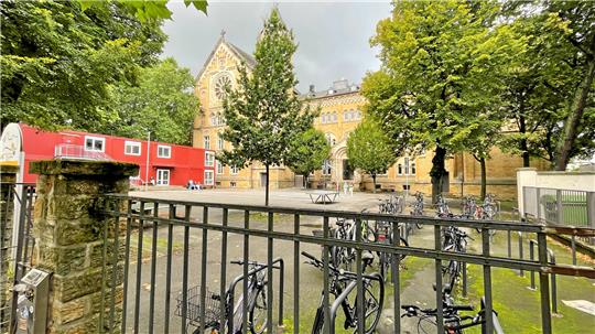 Blick durch ein schwarzes Metallgitter auf einen Innenhof mit mehreren Fahrrädern, Bäumen und einem historischen Backsteingebäude im Hintergrund