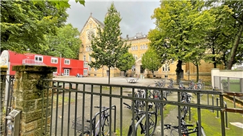 Blick durch ein schwarzes Metallgitter auf einen Innenhof mit mehreren Fahrrädern, Bäumen und einem historischen Backsteingebäude im Hintergrund