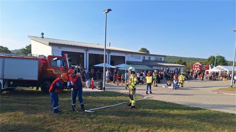 Feuerwehrleute und Jugendwehr-Mitglieder in Schutzkleidung bei einer Übung vor einem Feuerwehrhaus mit Einsatzfahrzeug und Zuschauern im Hintergrund