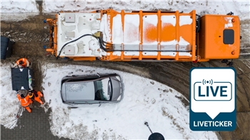 Eine Luftaufnahme: Mitarbeiter der Müllabfuhr schieben eine große Mülltonne in Richtung eines Müllwagens. 