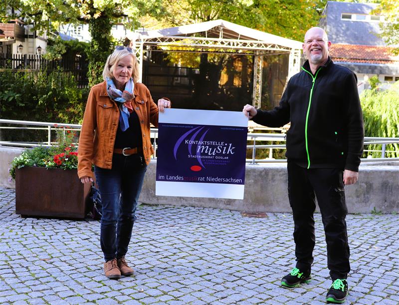 Zwei Menschen, eine Frau und ein Mann, stehen auf einem gepflasterten Boden vor einer Bühne und halten ein Plakat hoch.