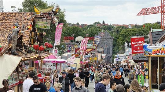 Menschenmenge auf einem belebten Jahrmarkt mit bunten Ständen, darunter ein Stand mit großen Erdbeer-Dekorationen und ein Stand mit einem Schild 'Kartoffelpuffer & Pommes'