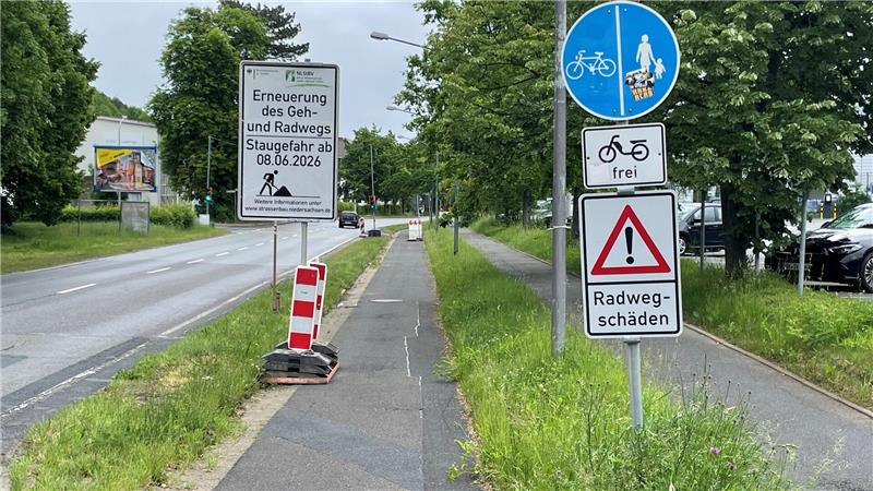 Radweg mit mehreren Verkehrsschildern, darunter ein blaues Schild für Radfahrer und Fußgänger, ein Schild mit der Aufschrift 'frei', ein Warnschild mit Ausrufezeichen und dem Text 'Radwegschäden' sowie ein Baustellenschild mit dem Hinweis auf Erneuerung des Geh- und Radwegs bis 8.6.2026.