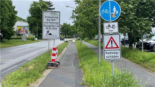 Radweg mit mehreren Verkehrsschildern, darunter ein blaues Schild für Radfahrer und Fußgänger, ein Schild mit der Aufschrift 'frei', ein Warnschild mit Ausrufezeichen und dem Text 'Radwegschäden' sowie ein Baustellenschild mit dem Hinweis auf Erneuerung des Geh- und Radwegs bis 8.6.2026.