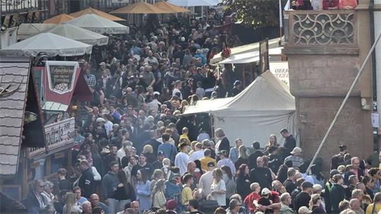 Menschenmenge auf einer belebten Straße mit Fachwerkhäusern und Marktständen an einem sonnigen Tag