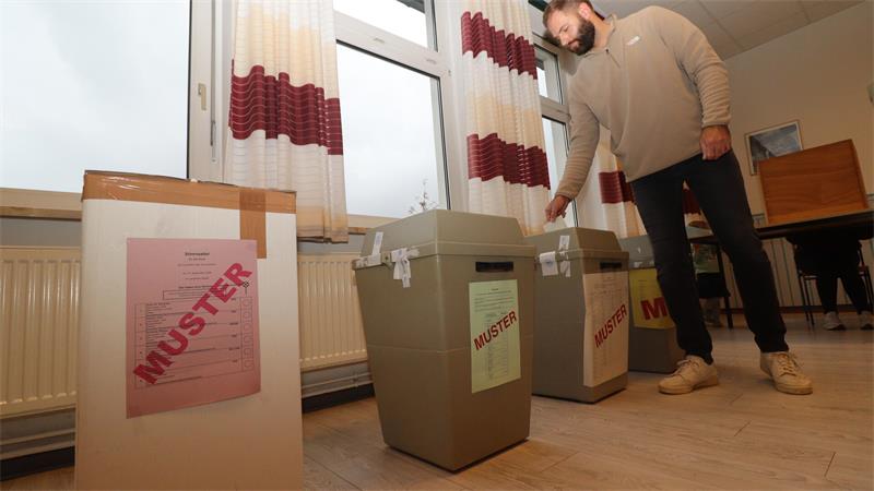 Mann wirft Zettel in graue Wahlurne in einem Raum mit Fenstern und rot-weißen Vorhängen.