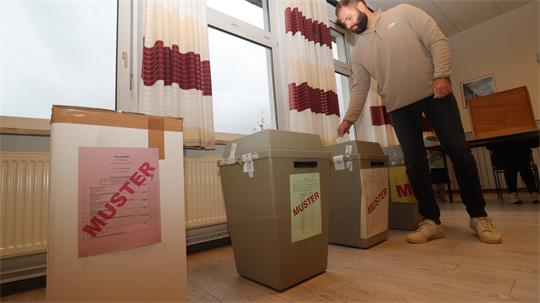 Mann wirft Zettel in graue Wahlurne in einem Raum mit Fenstern und rot-weißen Vorhängen.