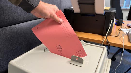 Jemand schiebt einen roten Wahlzettel in den Schlitz einer Wahlurne.