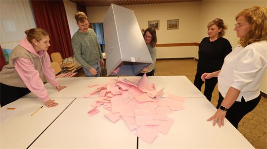 Fünf Personen stehen um einen Tisch, während zwei von ihnen einen großen Behälter mit vielen rosa Zetteln darauf kippen.