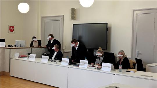 Mehrere Personen mit Mund-Nasen-Schutz sitzen an einem langen Tisch in einem Konferenzraum mit großem Bildschirm und geschlossener Tür im Hintergrund