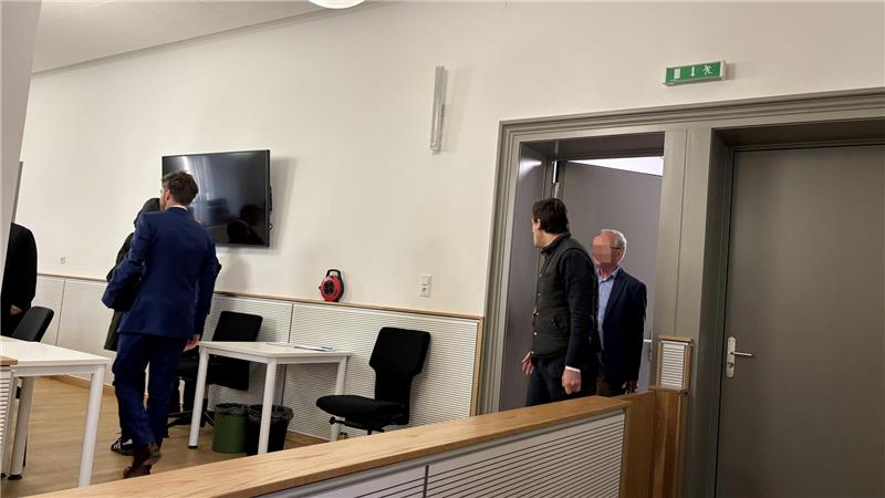 Mehrere Männer in Anzügen und Jacken gehen durch einen hellen Raum mit Stühlen, einem Fernseher an der Wand und einem offenen Türrahmen.