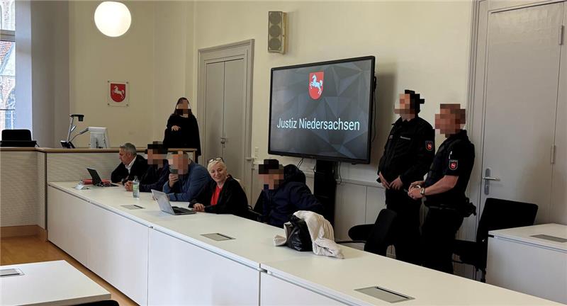 Ein Gerichtssaal. An einem Tisch sitzen Personen. Dahinter stehen Wachleute. 