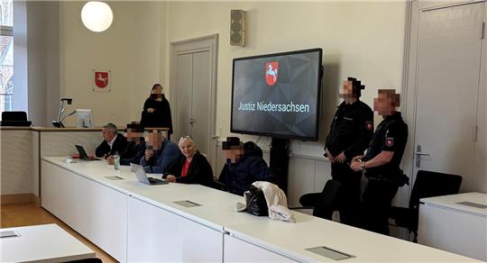 Ein Gerichtssaal. An einem Tisch sitzen Personen. Dahinter stehen Wachleute. 