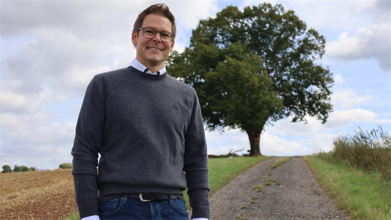 Ein lächelnder Mann mit Brille, grauem Pullover und weißem Hemd steht auf einem Feldweg vor einem großen Baum unter bewölktem Himmel.