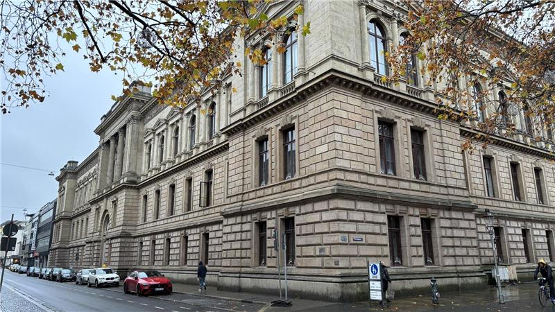 Das Braunschweiger Landgericht: ein großes historisches Gebäude mit Sandsteinfassade an einer nassen Straße, mehrere parkende Autos und Fußgänger, herbstliche Bäume mit gelben Blättern.