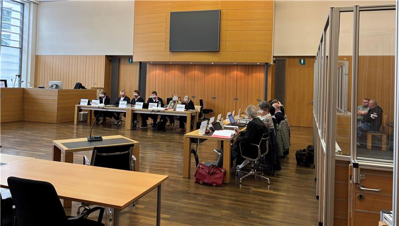 Menschen sitzen an Tischen in einem großen Raum mit Holzverkleidung und einem Bildschirm an der Wand.