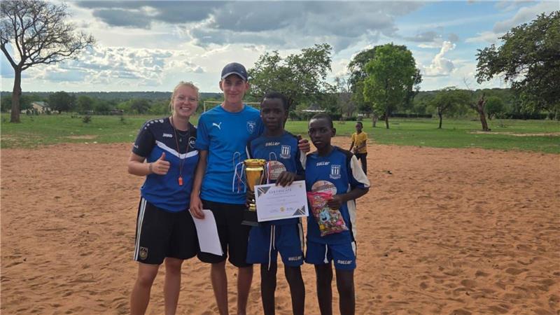 Goslarerin Amalia Saupe trainiert Mädchenteam in Afrika (Teil 2) Vier Personen stehen auf einem sandigen Spielfeld, drei davon tragen blaue Sportkleidung, eine Person hält eine Urkunde und eine Trophäe, eine weitere eine Tüte mit Snacks.