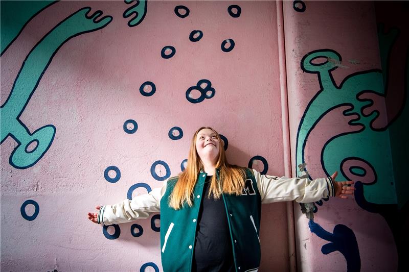 Studium mit Trisomie 21: Amelie Gerdes (18) macht es vor Amelie Gerdes steht an einer bunt bemalten Wand vor ihrem Tanzstudio. Die junge Bremerin mit Trisomie 21 studiert seit kurzem zusammen mit drei weiteren jungen Menschen mit Lernschwierigkeiten an der Hochschule für Künste im Sozialen (HKS) in Ottersberg. Foto: Sina Schuldt/picture alliance/dpa