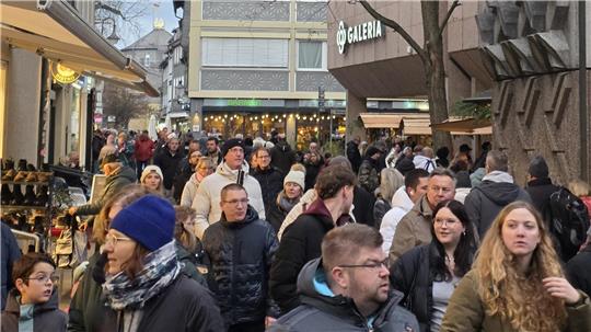 Menschenmenge auf einer belebten Einkaufsstraße mit Geschäften und Bäumen, im Hintergrund ein Gebäude mit dem Schriftzug 'GALERIA'.