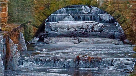 Gefrorener Fluss unter einem steinernen Bogen mit Eisschichten und gefrorenem Wasser.