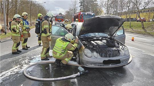 Mehrere Feuerwehrleute in Schutzkleidung stehen um ein Auto mit geöffneter Motorhaube auf einer Straße, aus der Flüssigkeit austritt