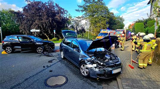 Zwei Autos mit beschädigten Frontschürzen stehen auf einer Kreuzung. Auf dem Gehsteig stehen Feuerwehrleute, die zum Teil Besen in den Händen halten.