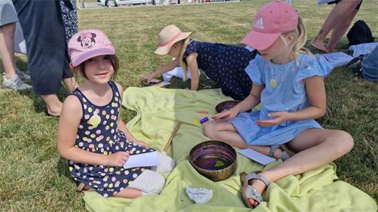 Zwei Mädchen mit Mützen sitzen auf einer Decke im Gras und spielen mit kleinen Gegenständen.