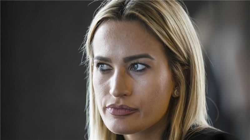 Frau mit langen blonden Haaren trägt schwarzen Blazer und sitzt vor dunklem Hintergrund.