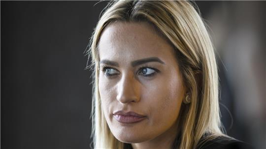 Frau mit langen blonden Haaren trägt schwarzen Blazer und sitzt vor dunklem Hintergrund.