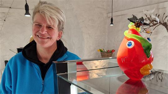 Lächelnde Person mit grauem Haar und blauem Oberteil neben einer bunten, vogelähnlichen Keramikfigur auf einem Glasregal
