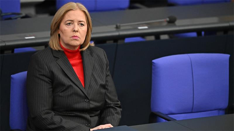 Eine blonde Frau mit kurzen Haaren und einem schwarzen Anzug sitzt auf einem blauen Stuhl an einem Tisch.