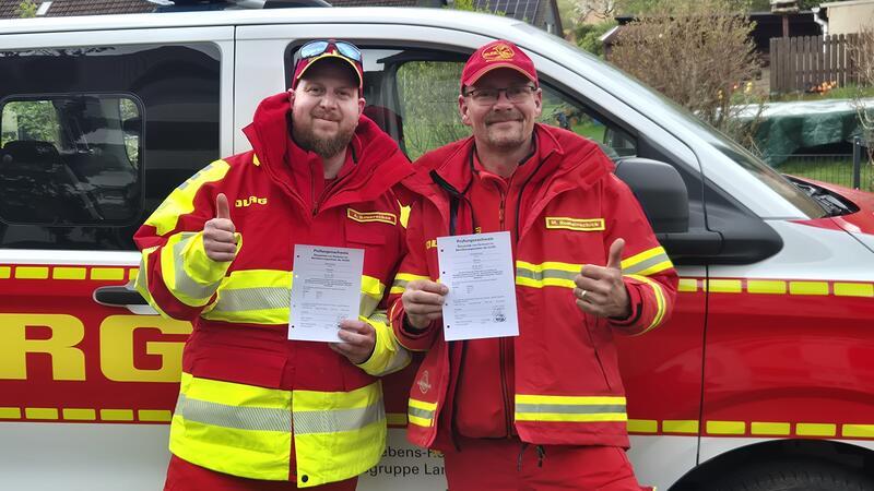 Zwei Personen in rot-gelber Einsatzkleidung halten jeweils ein Dokument und zeigen Daumen nach oben vor einem Einsatzfahrzeug mit roter und gelber Beschriftung