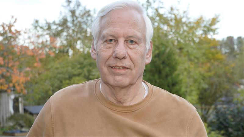Älterer Mann mit weißem Haar und braunem Pullover vor herbstlicher Baumkulisse.