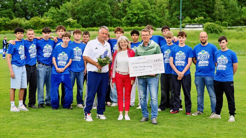 Gruppe von Jugendlichen und Erwachsenen auf einem Sportplatz, zwei Personen halten einen großen Scheck und eine Person einen Blumenstrauß.