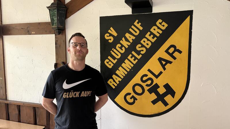 Person mit schwarzem T-Shirt mit Aufdruck 'GLÜCK AUF' steht vor einer Wand mit großem gelb-schwarzem Wappenschild mit der Aufschrift 'SV GLÜCKAUF RAMMELSBERG GOSLAR' und einem Bergmannssymbol