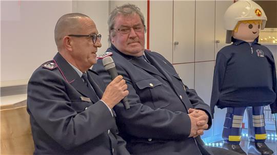 Zwei Männer in dunklen Uniformen sitzen nebeneinander, einer hält ein Mikrofon. Daneben steht eine große Playmobilfigur in Feuerwehruniform mit Helm.