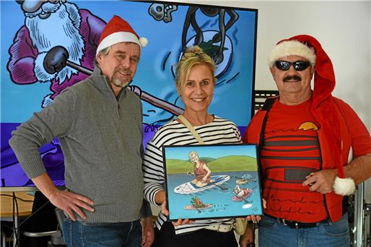 Anette von Burgdorf freut sich über ihren eigenen Sup-Cartoon von Thomas Velte (li.), der die Ausstellung „Auch der Weihnachtsmann fährt SUP“ mit Günter Koschig (re.) im Mach-Mit-Haus initiiert hat.