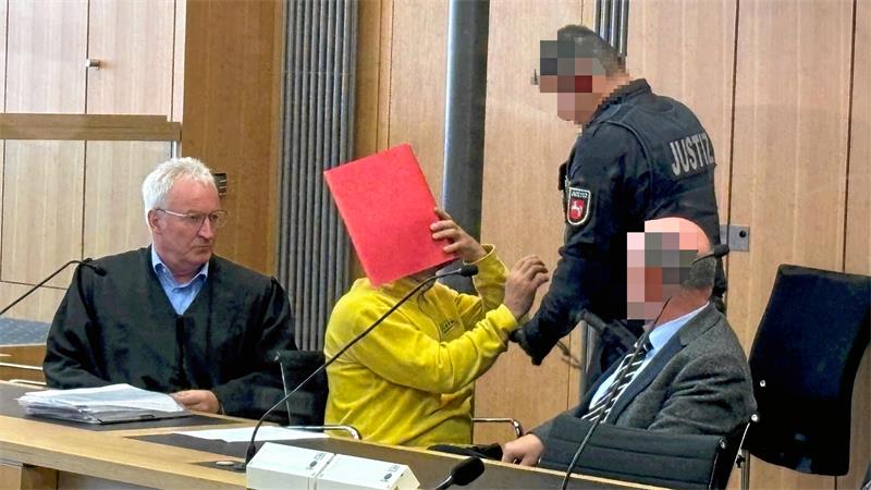 Person in gelber Kleidung hält rotes Dokument vor Gesicht, sitzt neben zwei Männern in Anzügen, ein Justizbeamter steht daneben in einem Raum mit Holzvertäfelung