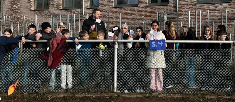 Goslarer CvD-Gymnasium erläuft 8500 Euro für „Girls in Action“ Gruppe von Kindern und Erwachsenen steht hinter einem Maschendrahtzaun, ein Mädchen hält ein Schild mit der Zahl '5.4'