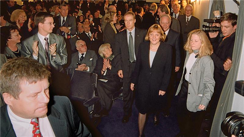 Menschen in formeller Kleidung in einem Raum, einige applaudieren, andere stehen oder sitzen