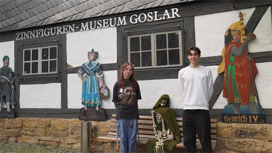 Gruppenfoto vor dem Zinnfiguren-Museum Goslar. Zwei junge Personen stehen vor dem historischen Fachwerkgebäude neben einer Bank mit einem als Skelett dargestellten Ausstellungsobjekt. An der Fassade sind historische Figuren sowie die Beschriftung „Zinnfiguren-Museum Goslar“ und eine Darstellung Heinrichs IV. zu sehen.