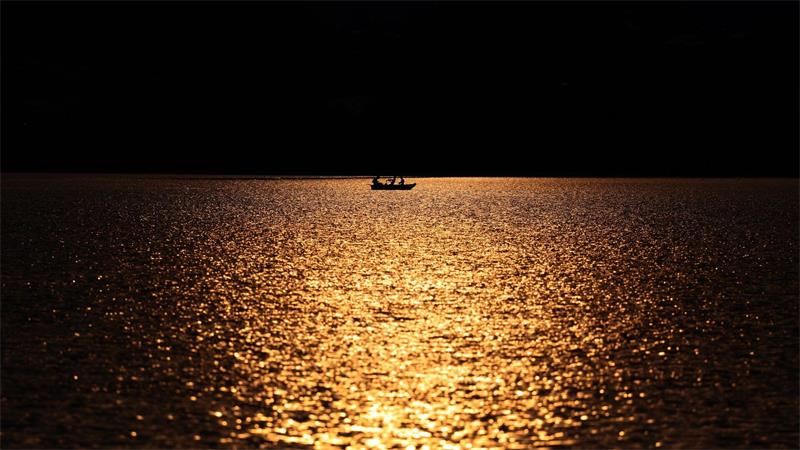 Angler rudern in einem Kahn über den im Sonnenuntergang glitzernden Hopfensee.