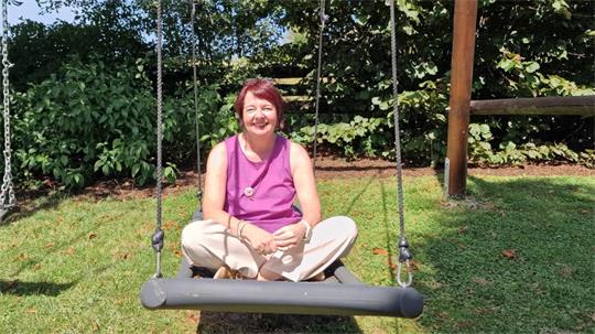 Frau sitzt im Schneidersitz auf einer Schaukel im Garten bei Sonnenschein.
