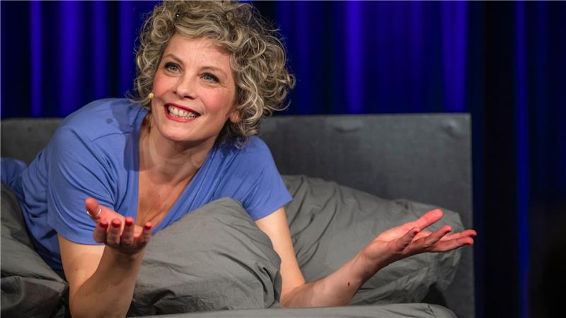 Eine hübsche Blondine mit Locken liegt bäuchlings auf einer Bettdecke und streckt lächelnd die Hände in die Luft.