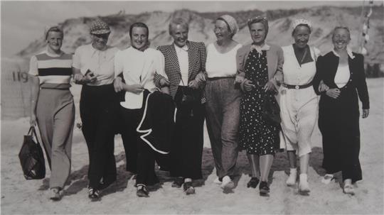 Sieben Personen stehen nebeneinander im Freien, einige tragen Hüte und unterschiedliche Freizeitkleidung.